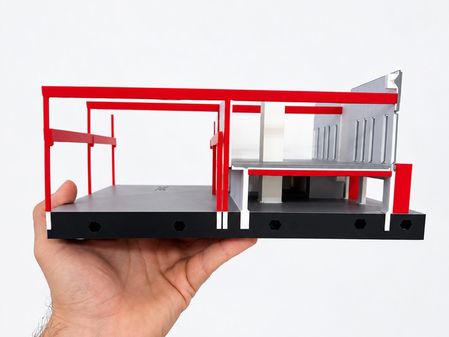 One printed section held in a hand, cut to show the mezzanine level, red beams and dowel holes in the base